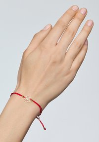 Czerwony pleciony sznurkowy bransoletka z złotym charms w kształcie kwiatu i małymi złotymi koralikami, prezentowana na dłoni na neutralnym tle.