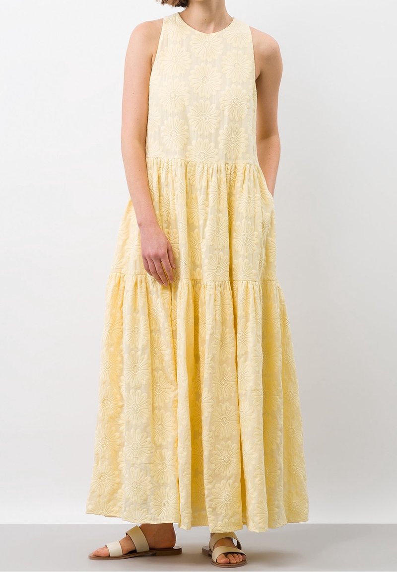 Femme portant une robe maxi sans manches, jaune, brodée de fleurs avec des poches, se tenant contre un fond neutre.