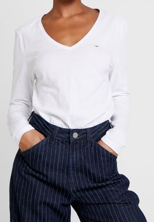 Osoba nosící bílou dlouhý rukáv s výstřihem do V, zastrčenou do tmavě modrých pruhovaných vysokopásových kalhot, s rukama v kapsách.