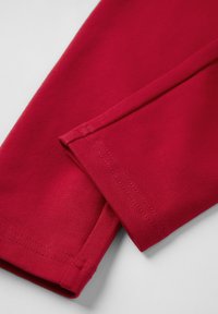 Punainen puuvillakangas, jossa on ressattuja hihoja, esittelee sileää pinta-alaa ja selkeitä ompeleita. Suunnittelussa on yksinkertainen ja siisti leikkaus.