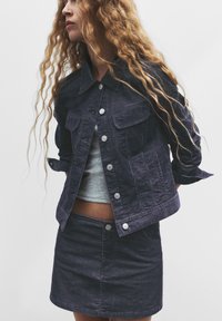 Jachetă din denim închis cu butoane argintii și buzunare pe piept, asociată cu o fustă din denim închis. Ținuta include un top gri deschis dedesubt.