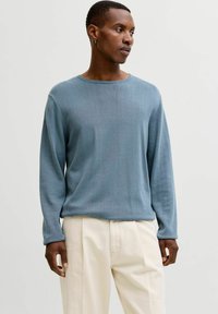 Homme portant un pull à manches longues bleu clair et un pantalon de couleur crème, regardant vers sa droite sur un fond blanc uni.