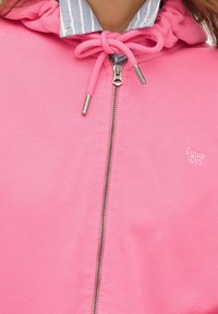 Close-up van een roze zip-up hoodie met een gestrikte trekkoord en een gestreepte shirtkraag zichtbaar eronder.