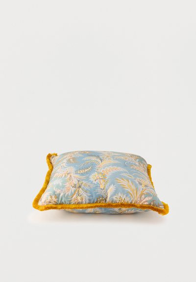 Coussin décoratif carré avec motif floral bleu et bordure à franges jaunes, posé sur une surface blanche.
