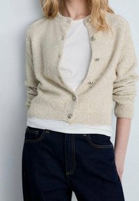 Beige neulottu neuletakki, jossa on teksturoitu kuvio, pyöreä pääntie, hopeanväriset napit, päällä valkoinen paita ja yhdistetty tummien farkkujen kanssa.