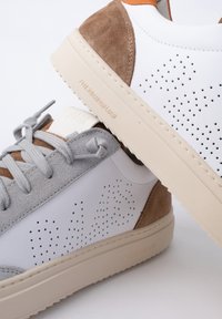 Zapatillas blancas con acentos de ante gris y marrón, diseño perforado, suela plana beige y cordones texturizados. Marca en el lateral.