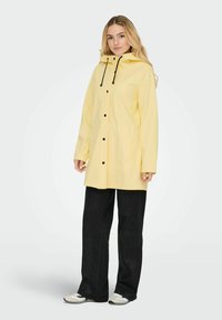 Rumena vodoodporna jakna s kapuco, sprednjimi gumbi in črnimi vrvicami; kombinirana s črnimi širokimi hlačami in nevtralnimi čevlji.