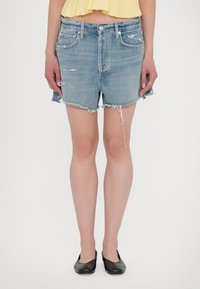Hellblaue, hochgeschneiderte Jeansshorts mit fransigem Saum, ausgestattet mit einem verwaschenen Design und klassischen Taschen. Kombiniert mit schwarzen Ballerinas.