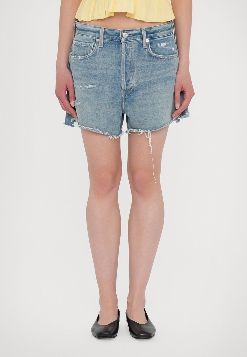 Hellblaue, hochgeschneiderte Jeansshorts mit fransigem Saum, ausgestattet mit einem verwaschenen Design und klassischen Taschen. Kombiniert mit schwarzen Ballerinas.