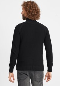 Maglione nero lavorato a maglia con colletto alto e polsini a coste, caratterizzato da un motivo testurizzato. Dettagli di cucitura visibili sulle spalle e sull'orlo.