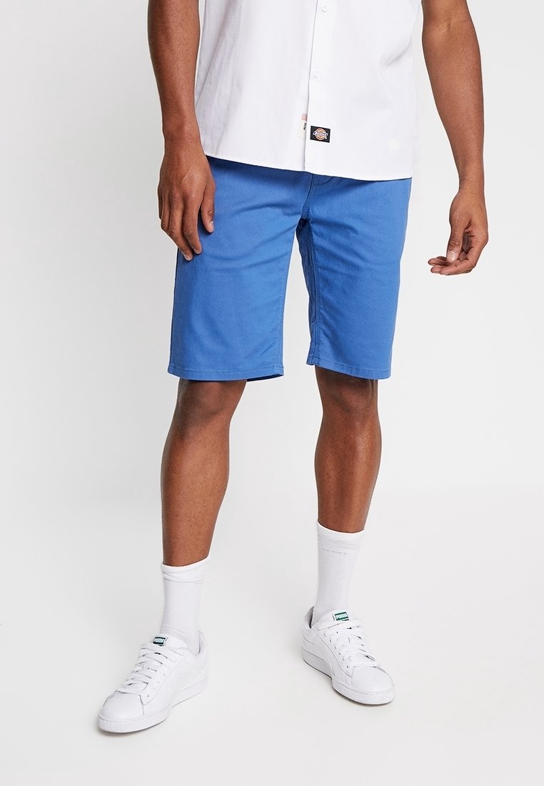 Homme portant un short bleu vif, une chemise blanche, des chaussettes blanches et des baskets blanches, debout devant un fond uni.