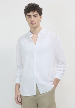 Long-sleeve, white button-up shirt made of lightweight fabric, featuring a classic collar and a relaxed fit, paired with beige pants.