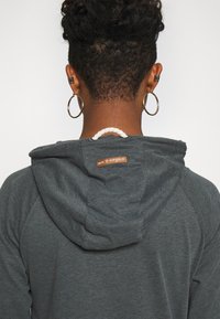 Grijze hoodie met een hoody met trekkoord, voorzien van een bruine leren label met de tekst "we ❤️ vegan." Zacht materiaal en raglanmouwen zichtbaar.
