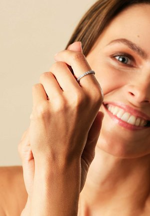 Nahaufnahme einer lächelnden Frau, die einen mit Diamanten besetzten Ring an ihrer linken Hand vor einem schlichten beige Hintergrund zeigt.