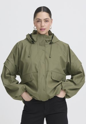 Frau mit dunklem Haar in einem Dutt, trägt Creolen, eine olivgrüne Kapuzenjacke mit Taschen und schwarze Hose, Hände in den Jackentaschen.