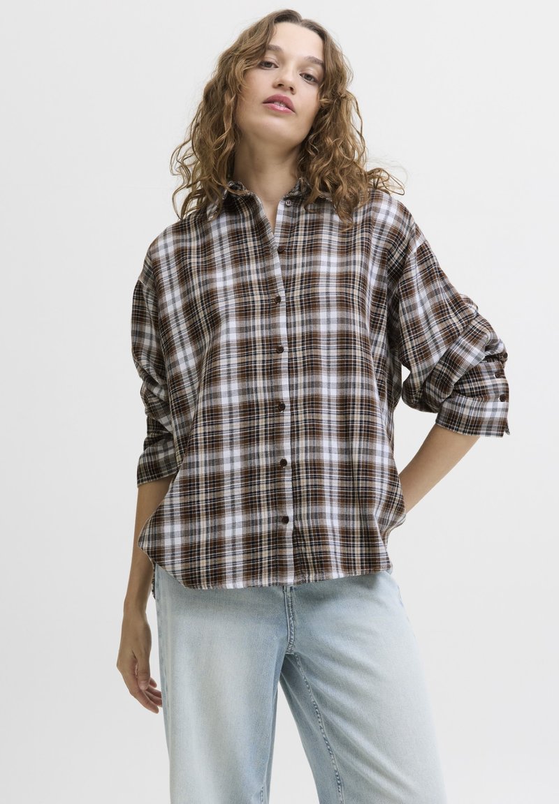 Kvinde stående med hånden på hoften iført brun og hvid ternet skjorte med knapper og lyseblå jeans mod en ensfarvet baggrund.