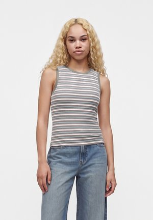 Jeune femme aux longs cheveux blonds et bouclés, portant un haut rayé sans manches et un jean bleu, debout devant un fond uni.