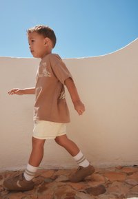 Niño pequeño caminando al aire libre sobre un pavimento de piedra, vestido con una camisa marrón, pantalones cortos blancos, calcetines a rayas y zapatos marrones frente a una pared beige y un cielo azul.