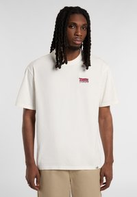 Camiseta de algodón color crema con mangas cortas y un logotipo bordado rojo en el lado izquierdo del pecho. Corte recto y ajuste relajado.