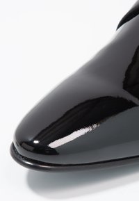 Chaussure en cuir verni noir avec un bout pointu et une surface brillante, présentant une texture lisse et un design minimaliste.