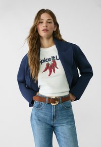 Blazer marine sur un t-shirt blanc avec un graphique de piment rouge, associé à un jean bleu et une ceinture marron avec une boucle dorée. Tenue simple et décontractée.