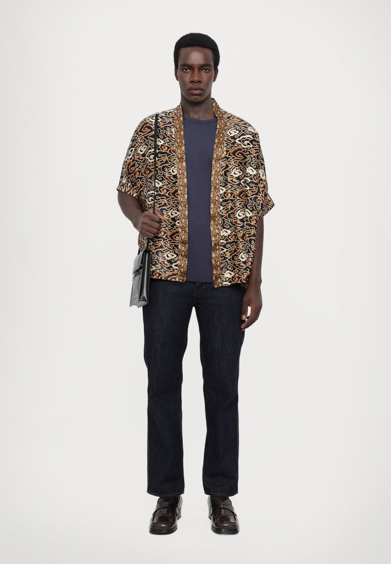 Košile s krátkým rukávem s květinovým vzorem v hnědé, krémové a zlaté barvě, spárovaná s tmavě modrými džíny a černými koženými botami. Černá taška přes rameno.