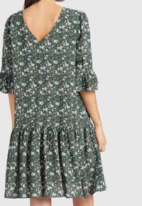 Vestido floral verde con escote en V, mangas acampanadas y dobladillo con volantes. Presenta un patrón de flores blancas y hojas verdes sobre un fondo oscuro.