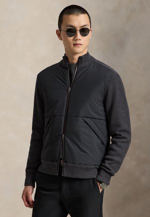 Homme portant des lunettes de soleil noires et une veste noire zippée avec des manches tricotées, posant avec une main dans la poche devant un fond neutre.