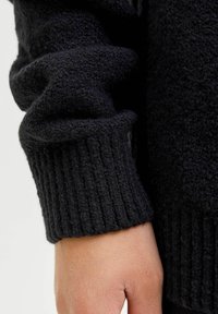 Mustva, tekstureeritud sviiteris alongu ribidega; pehme, karvane kangas loob visuaalse kontrasti paljastatud käe siledaga.