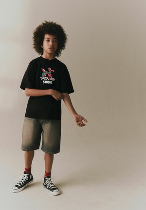 Barn med krøllet hår iført en sort grafisk t-shirt, grå shorts, røde sokker og sorte høje sneakers, stående foran en ensfarvet baggrund.