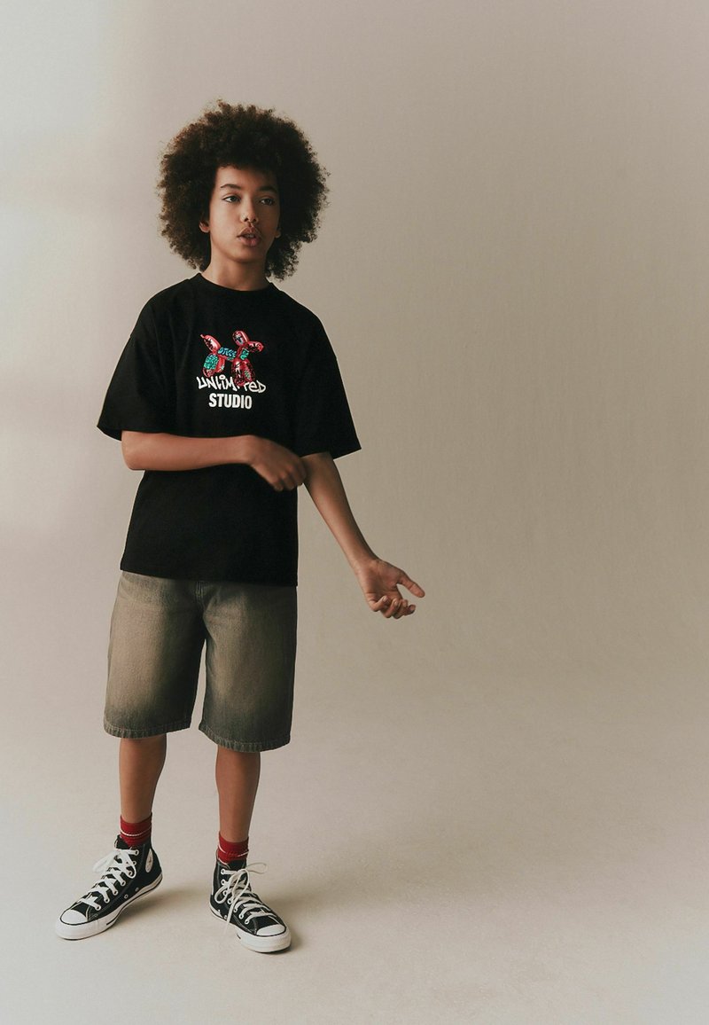 Göndör hajú gyermek, fekete grafikás pólóban, szürke rövidnadrágban, piros zokniban és fekete magasszárú tornacipőben, egyszerű háttér előtt állva.