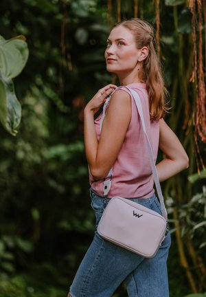 Crossbody bag in light pink, rectangular shape with smooth texture. Features a black logo and adjustable strap, worn by a model in a green setting.