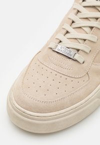 Béžový semišový špičatý svršek tenisky s perforacemi, béžové tkaničky a kovová známka na tkaničkách s vyrytým nápisem "Steve Madden".