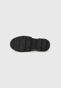 Semelle de chaussure en caoutchouc noir avec des rainures horizontales et un talon arrondi. Présente un logo en relief et une surface texturée pour une meilleure adhérence.