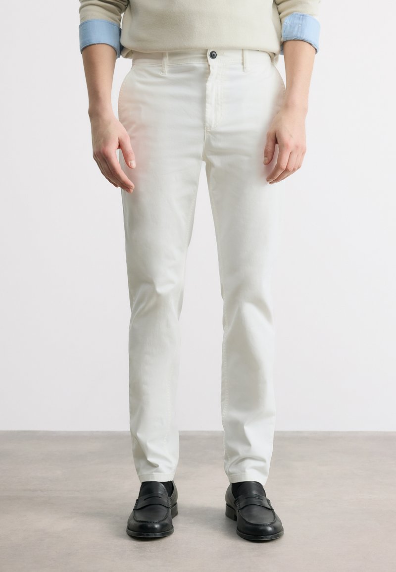 Homme portant un pantalon blanc droit, des mocassins noirs et un pull beige avec des manches bleues retroussées, debout sur un sol gris.