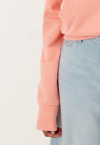 Personne portant un haut à manches longues rose avec des manches à poignets et un jean en denim bleu clair, avec un accent sur l'avant-bras et la taille.
