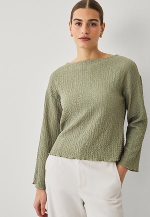 Kvinde med trukket hår tilbage iført guldringe i ørerne, en tekstureret salviegrøn langærmet top og hvide bukser med den ene hånd i lommen.