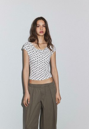 Mladá žena s dlhými hnedými vlasmi, oblečená v bielom topán s bodkami a voľných olivovo zelených nohaviciach, stojaca pred jednoduchou svetlou stenou.