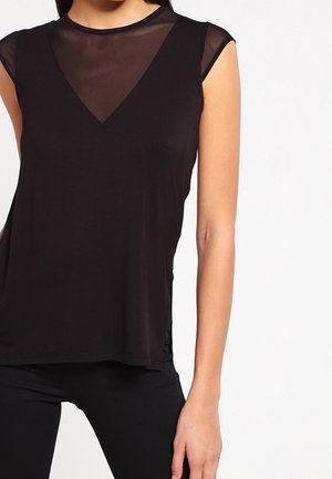 Vrouw draagt een zwarte top zonder mouwen met een transparante mesh inzet bij de halslijn, gecombineerd met een zwarte broek tegen een effen achtergrond.