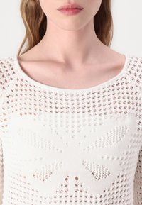 Hvit strikket genser med åpen vevd design og et sentralt sommerfugl mønster laget med større masker. Rund hals.