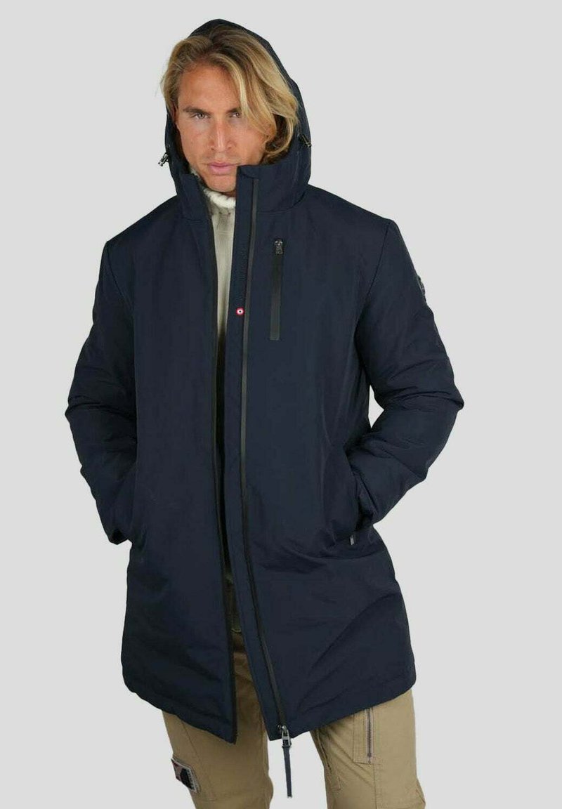 Homme aux cheveux blonds portant une veste d'hiver à capuche bleu marine et un pantalon beige, se tenant les mains dans les poches de la veste contre un fond uni.