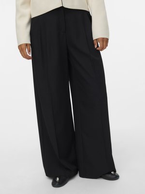 Άτομο που φοράει μαύρο παντελόνι με φαρδιά ποδόγυρα, ανοιχτό μπεζ τοπ και μαύρα μυτερά παπούτσια με μεταλλικές διακοσμήσεις, στέκεται μπροστά σε απλό φόντο.