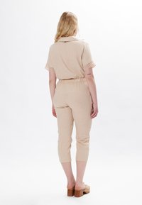 Jumpsuit in lino beige con maniche corte, vita elastica e gamba cropped. Presenta dettagli in crochet sulle maniche ed è abbinato a zoccoli con tacco in legno.