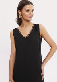 Czarna sukienka bez rękawów z dekoltem w kształcie litery V, zdobiona dekoracyjnym wykończeniem w formie pętli. Gładki materiał i luźny krój. Uzupełniona złotymi kolczykami.