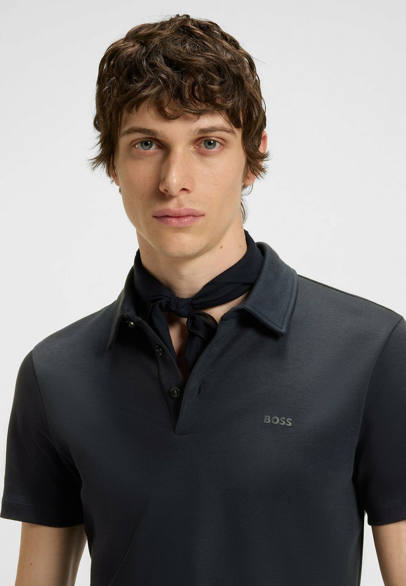 Sort polo skjorte med krave, prydet med et lille gråt "BOSS" logo. Modellen bærer et mørkt tørklæde bundet om halsen. Korte ærmer.