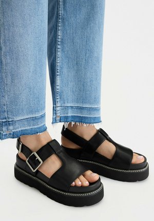 Pies con sandalias negras de plataforma de cuero con hebillas plateadas, combinadas con jeans de dobladillo deshilachado en azul claro sobre un fondo blanco.