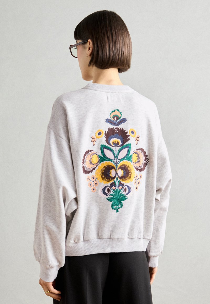 Šedá mikina s oversize strihom. Na zadnej strane má farebnú kvetinovú výšivku v žltej, zelenej, purpurovej a oranžovej farbe. Rybované manžety a lem.