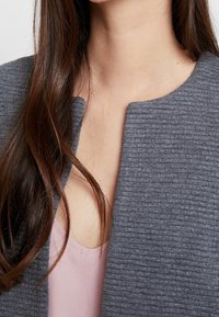 Chaqueta gris texturizada con un patrón suave y acanalado, superpuesta a una blusa de color rosa claro. Vista cercana del cuello y la tela.