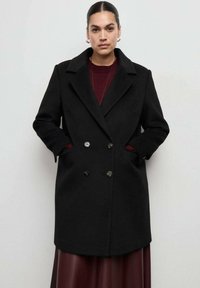 Czarny dwurzędowy płaszcz z wełny, z klapowym kołnierzem, bocznymi kieszeniami i ozdobnymi guzikami, zestawiony z bordowym swetrem pod spodem.