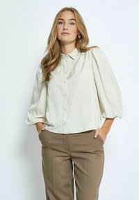 Light beige button-up blouse with a collared neckline, puffed sleeves, and a smooth texture, paired with high-waisted brown trousers.